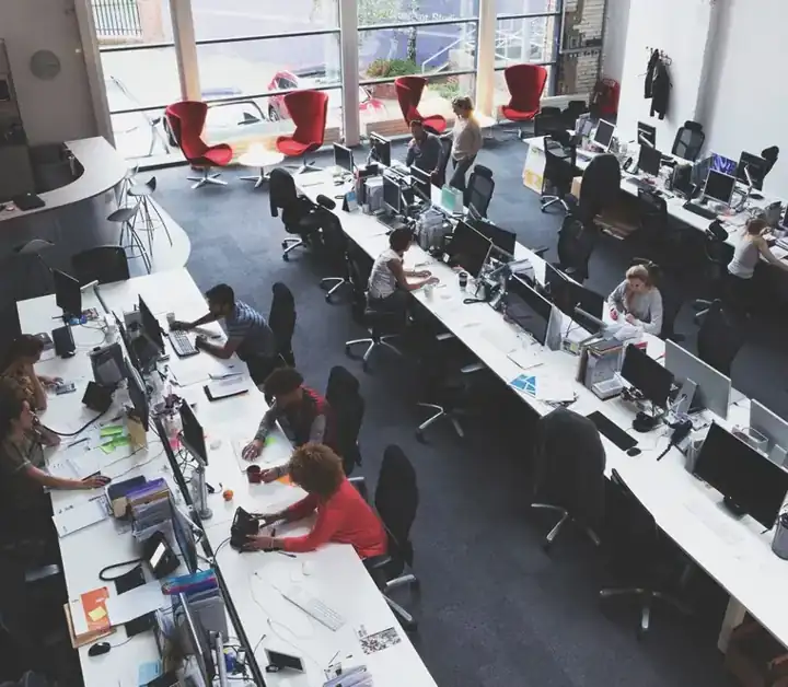 Open-plan office with long desks and multiple computer workstations; employees work at their computers amid large windows and scattered red lounge chairs in the background.