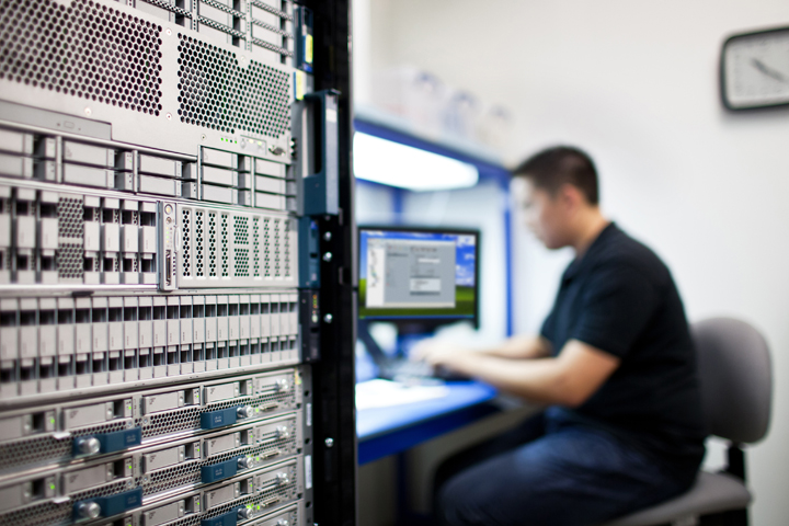 IT-Techniker sitzt an einem Arbeitsplatz neben einem hohen Server-Rack und überwacht einen Monitor in einer Rechenzentrumsumgebung.