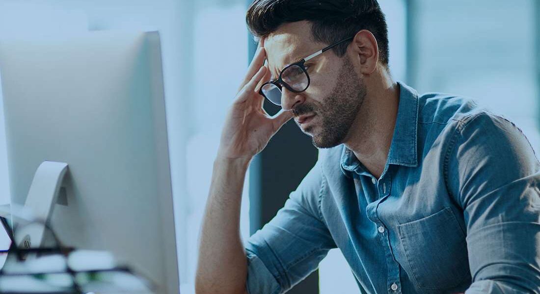Frustrated man in a denim shirt sits at a desk, hand resting on his forehead while looking at a computer screen.