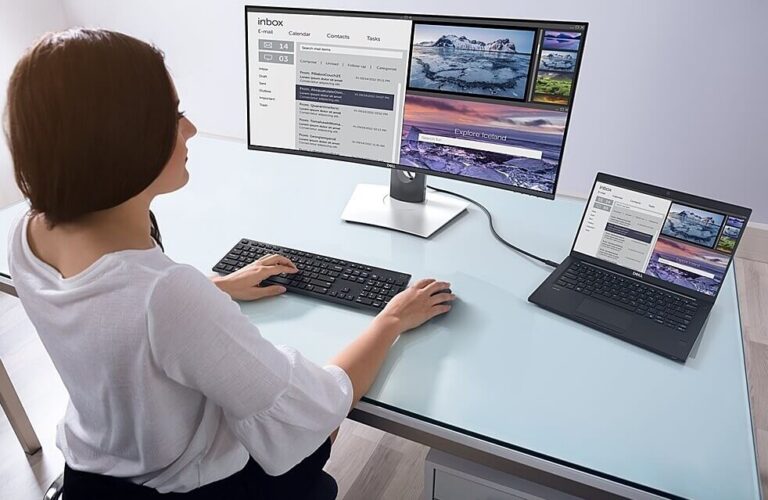 Personne utilisant un poste de travail à deux écrans : un grand moniteur et un ordinateur portable connecté sur un bureau en verre, affichant des e-mails et des photos côte à côte.
