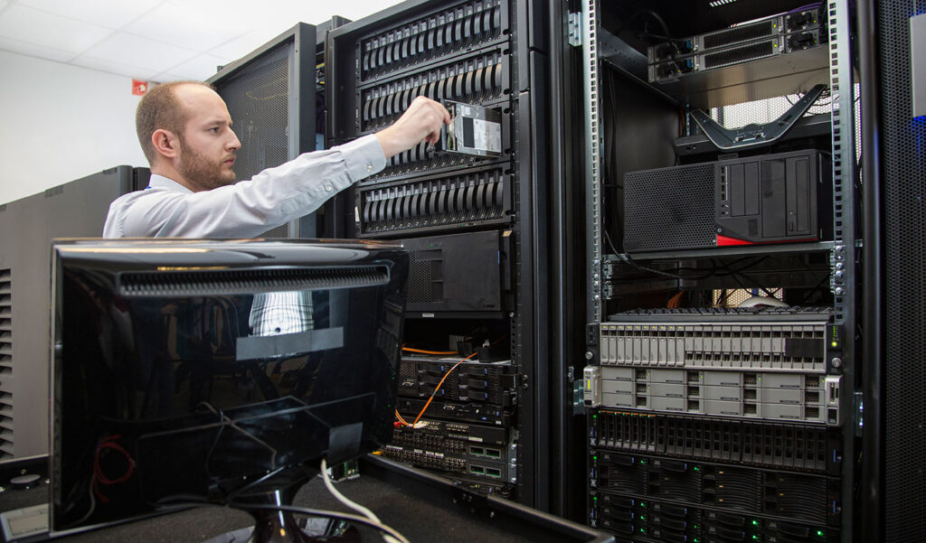 IT technician in a data centre inserts a hard drive into a server rack among multiple stacked servers and networking gear.
