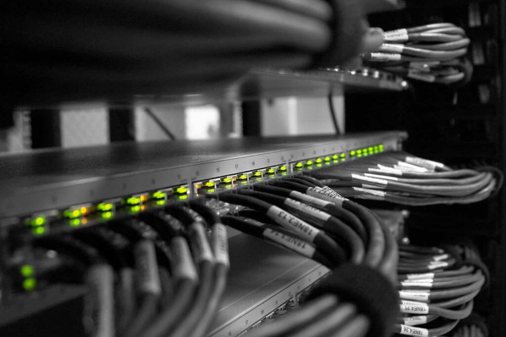 A network rack with a row of illuminated green LEDs on a switch and bundled Ethernet cables, labelled and organised inside a server cabinet