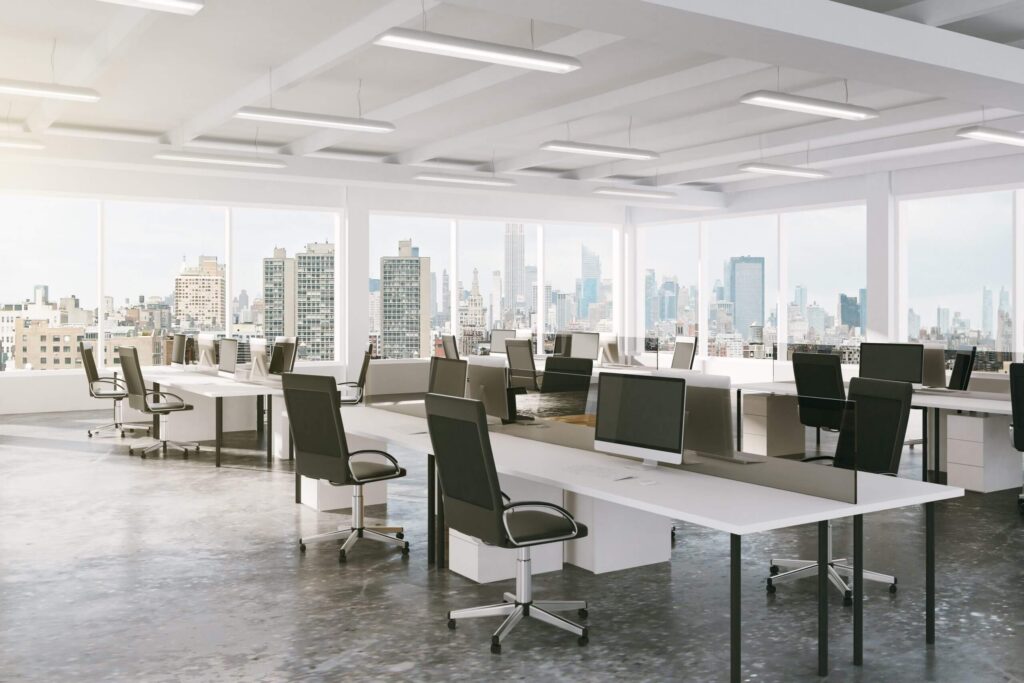 Bureau en open space avec des rangées de bureaux, des chaises ergonomiques et plusieurs écrans d'ordinateur, offrant une vue sur les toits de la ville à travers de grandes baies vitrées.