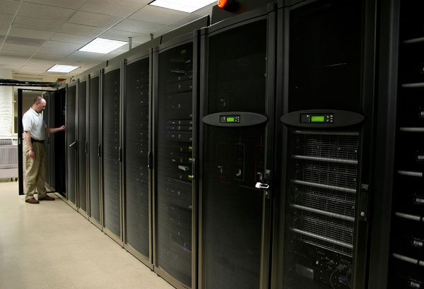 IT-Techniker im weißen Shirt steht an einer Reihe großer schwarzer Server-Racks in einem Rechenzentrumsflur, die Tür zu einem Schrank steht offen.