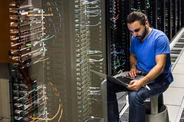 Tech-Mitarbeiter in einem blauen Hemd sitzt neben Server-Racks und benutzt in einem Rechenzentrum einen Laptop zur Fehlerbehebung.